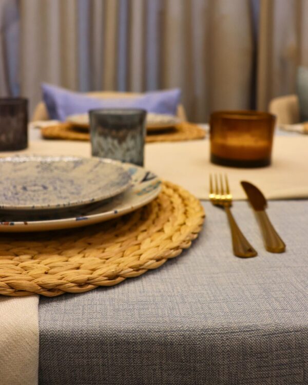 Detalle de mesa con mantel azul claro, cubiertos dorados, vaso texturizado y vela ámbar sobre camino de mesa beige.