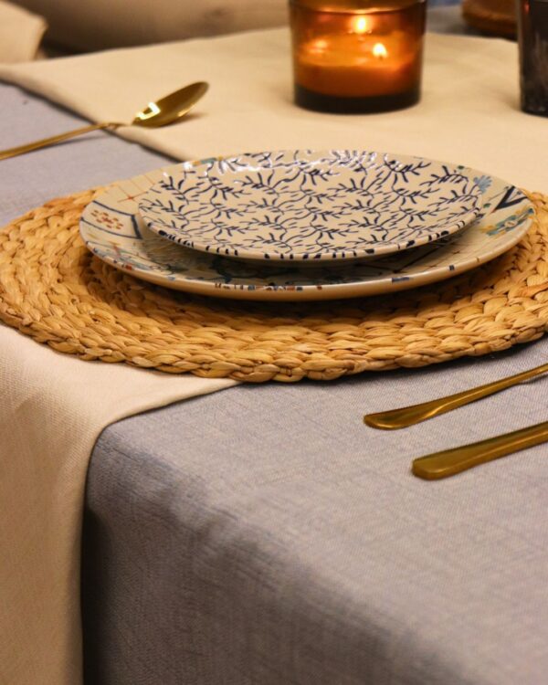Detalle de mesa con mantel azul claro, cubiertos dorados, vaso texturizado y vela ámbar sobre camino de mesa beige.